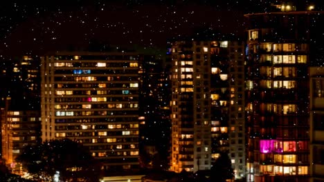 Snow-falling-against-view-of-tall-buildings-at-night-in-background