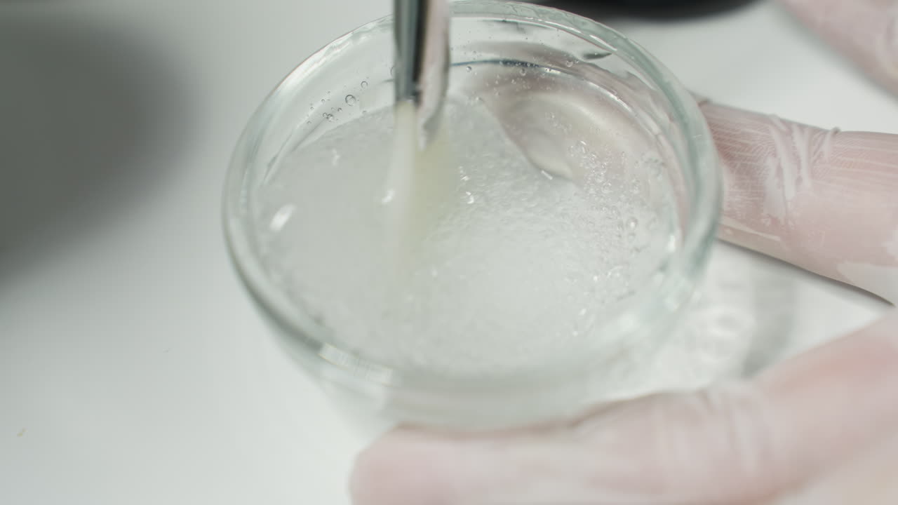 Protective Gloved Hand Stirring Face Scrub Gel In Glass Plate On White Tray Under Soft Light In Spa For Facial Treatment With Consistent Smooth Circular Motions To Enhance Skin Texture And Hydration