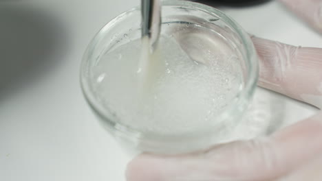 protective gloved hand stirring face scrub gel in glass plate on white tray under soft light in spa for facial treatment with consistent smooth circular motions to enhance skin texture and hydration