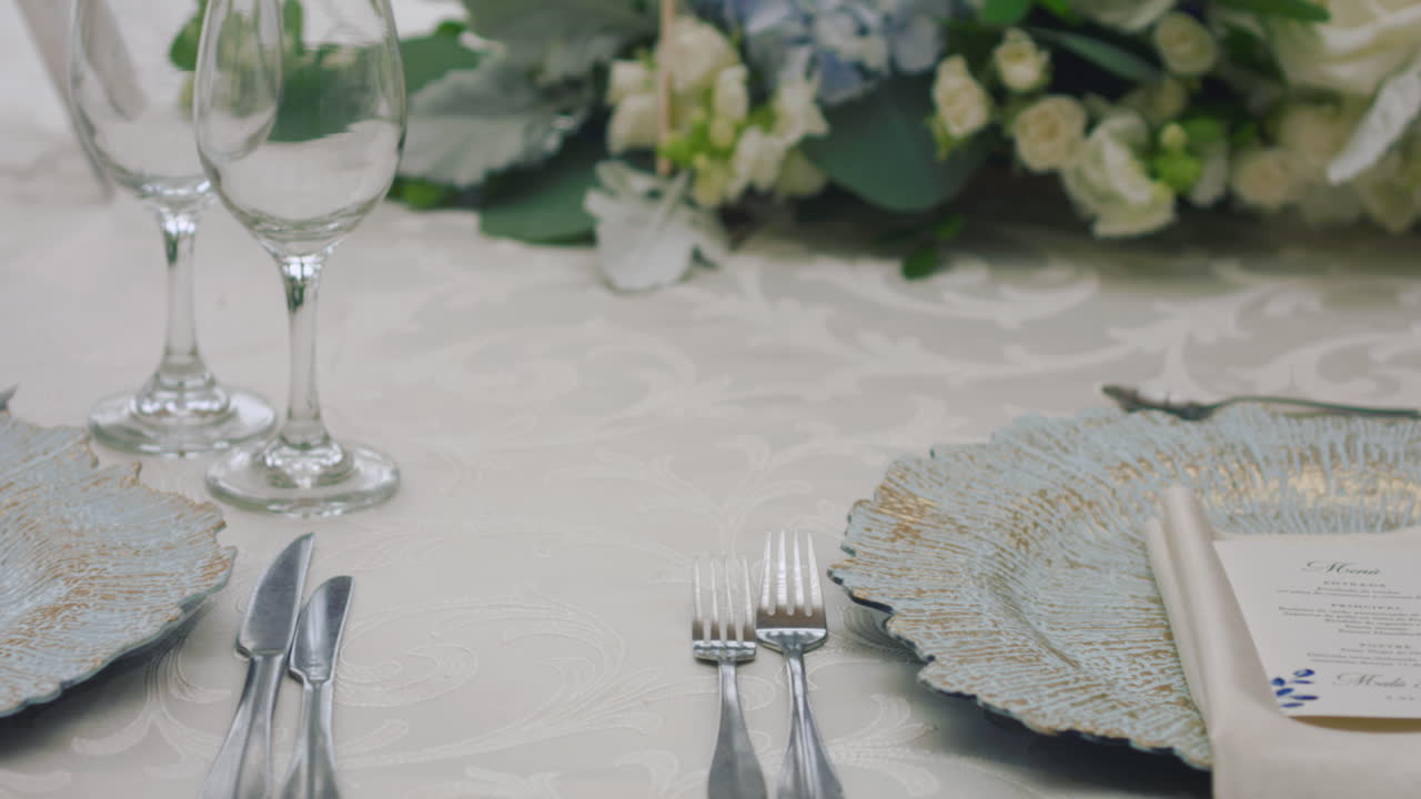 A detailed clip of an elegantly decorated wedding table. It features stemware, a white tablecloth, plates, cutlery, cloth napkins, a delicate floral arrangement, and a personalized invitation.