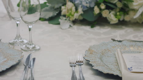 a detailed clip of an elegantly decorated wedding table. it features stemware, a white tablecloth, plates, cutlery, cloth napkins, a delicate floral arrangement, and a personalized invitation.