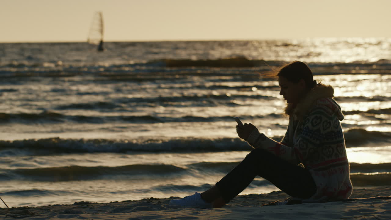 backgr에서 전화를 즐기는 모래에 앉아 바다 berugu에 젊은 여자의 실루엣