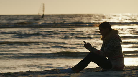 Silueta-De-Una-Mujer-Joven-En-El-Mar-Berugu-Sentado-En-La-Arena-Disfrutando-De-Un-Teléfono-En-El-Backgr