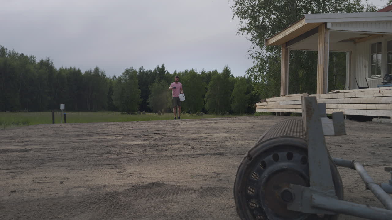 A man stands on a dirt ground with a house and forest in the background, with a piece of equipment in the foreground.