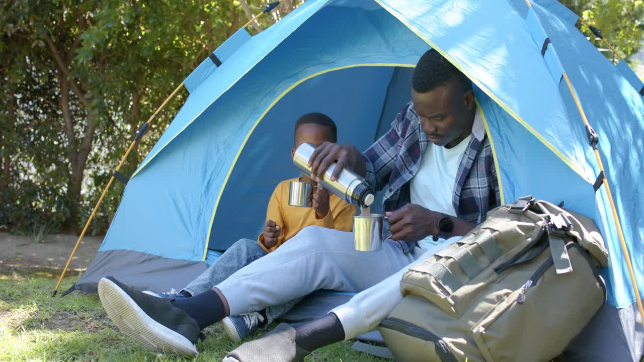 Afroamerikansk far og søn sidder ved teltindgangen, far rækker ud efter termoflasken og hælder op i kopper til at drikke.