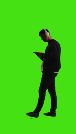 Vertical-Video-Full-Length-Shot-Of-Man-Wearing-Wireless-Headphones-Streaming-Music-From-Mobile-Phone-Against-Green-Screen-2