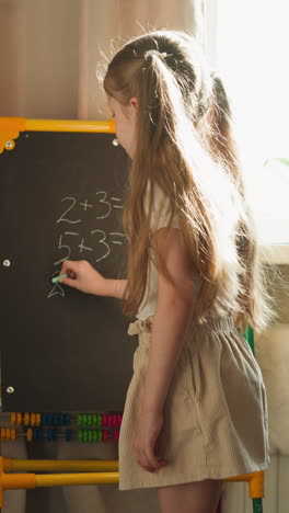 schoolgirl writes math problems on blackboard teaching younger brother slow motion. couple of children play school at home. siblings learn arithmetic together