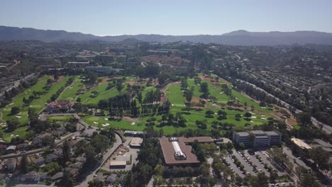 Aerial-helicopter-shot-of-golf-course-field-with-mountains-in-background-on-a-sunny-day-with-clear-blue-sky-pan-sideways