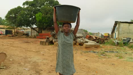 walking while carrying a water filled basin on top of her, a local woman is on her way back to her house in a neighborhood in kumasi, ghana, in africa