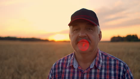 Retrato-De-Un-Granjero-Adulto-Mayor-En-Un-Campo-De-Grano-Mirando-La-Cámara-Y-Sonriendo-Al-Atardecer.-El-Conductor-Del-Tractor-Se-Quita-La-Gorra-Y-Mira-A-La-Cámara-En-Cámara-Lenta