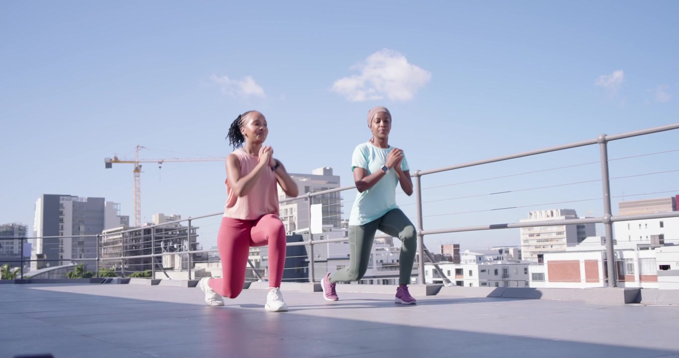 Two women starting workout, walking then lunging in sync on rooftop near dumbbells in workout gear