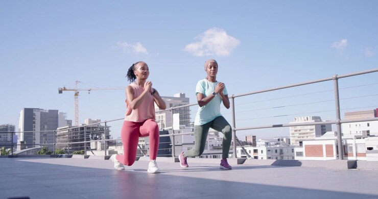 Two women starting workout, walking then lunging in sync on rooftop near dumbbells in workout gear