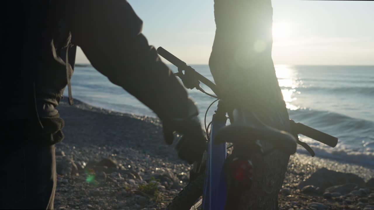 자전거를 타고 걷는 남자의 클로즈업, 나무 근처에서 쉬고 바위가 많은 해변에서 화창한 아침에 장갑을 벗다