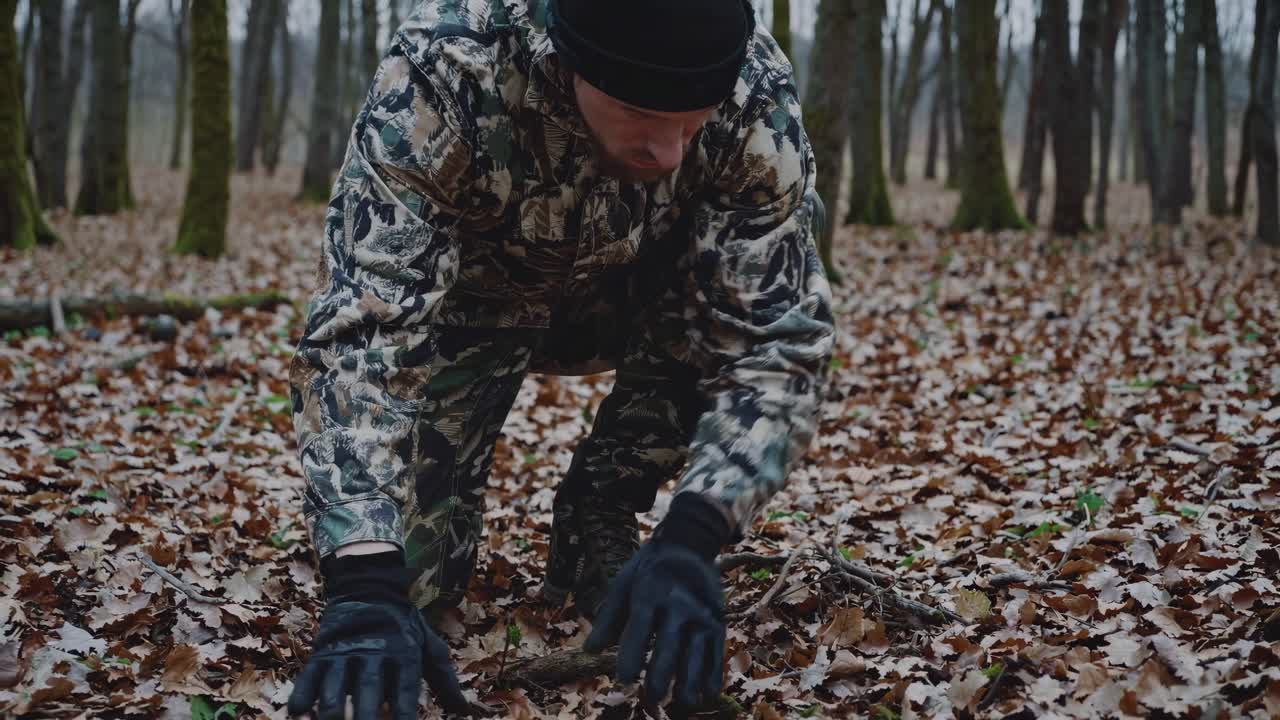Low-angle video shot of a person in camouflage gear kneeling in a forest, blending with fallen