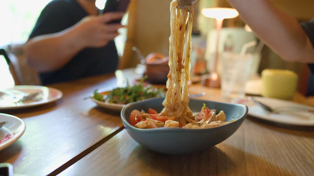 Person mixes spicy Thai noodle salad with utensils in bright, casual restaurant setting, shallow focus