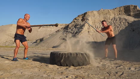 two male athletes training together hit the wheel with a hammer at sunset in the mountains on the sand. endurance training