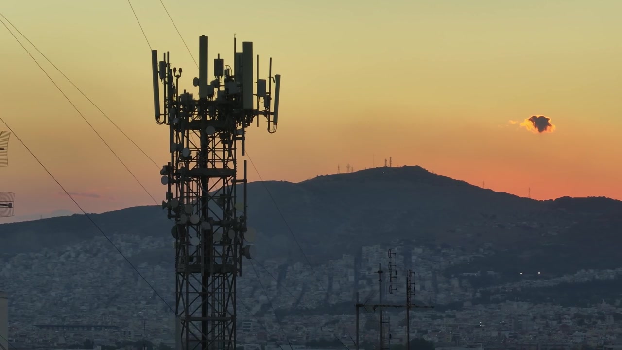 Drone orbit around communication antennas during sunset, revealing the glowing skyline of Athens and the sprawling cityscape beneath warm golden evening light.