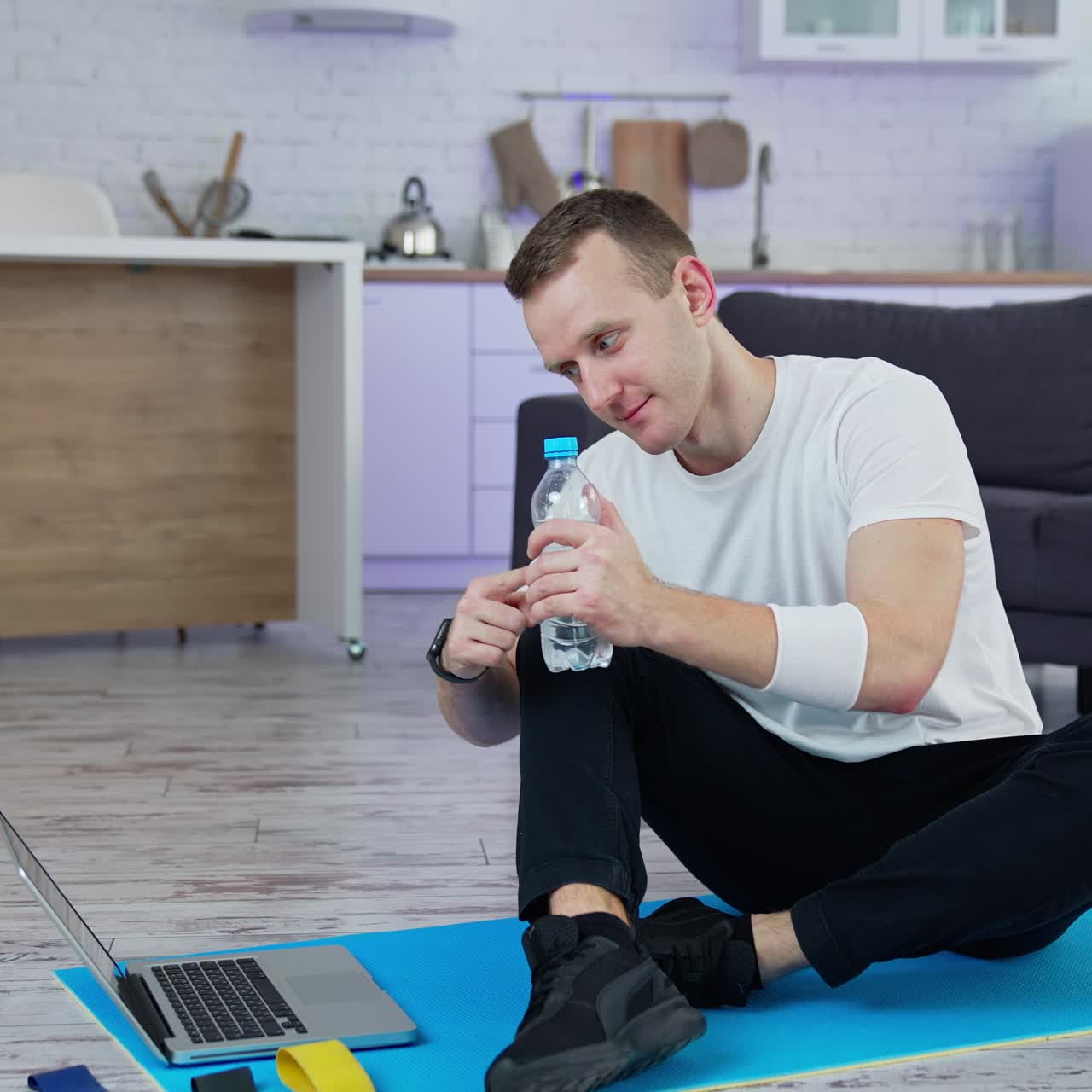 Young man sitting on a mat after training in front of a laptop. Sportive guy talking to someone through the laptop and shows a bottle of water in the kitchen