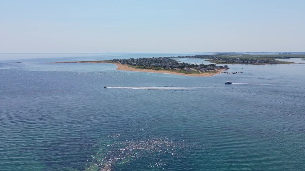 Barca con scia bianca che naviga attraverso un canale vicino a una lingua di sabbia che divide acque blu calme, con un campeggio per camper e pini in primo piano a Plum Island Beach, Massachusetts. Ripresa aerea con drone in sorvolo
