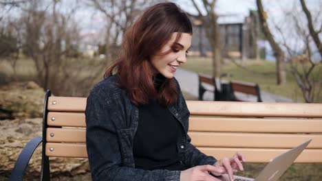 Attractive-female-in-her-20'sis-sitting-on-the-bench-at-park-typing-something-on-her-laptop-keyboard.-Enjoying-free-time.-Outdoors-footage