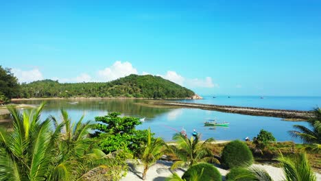Ruhiger-Ferienresortgarten-Mit-Palmen-Und-Dekorativen-Bäumen-Auf-Weißem-Sand,-Umspült-Von-Einer-Ruhigen-Lagune-Mit-Schwimmenden-Fischerbooten-In-Thailand