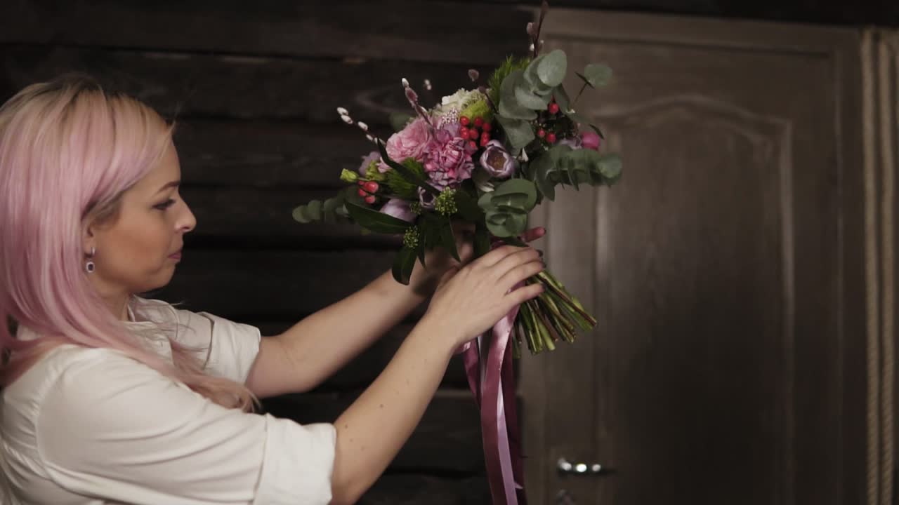A girl with pink hair holds a variety of flowers in her hands in front of her. The bouquet is decorated with dangling silk ribbons. Side view