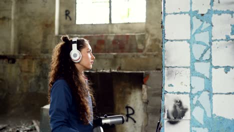 una mujer concentrada está decorando una vieja columna dañada dentro de un edificio industrial abandonado con graffiti usando pintura en aerosol y escuchando música con auriculares.
