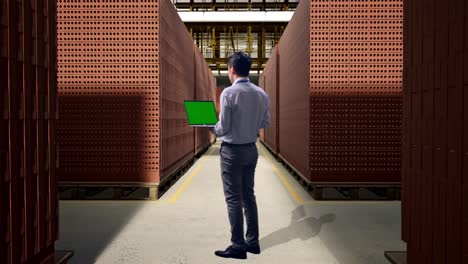 full body back view of an asian male professional worker standing with red brick packed in stacks are stored, typing on laptop's keyboard with green screen and look at the factory once
