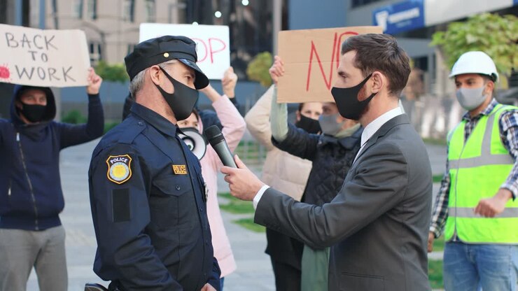 Caucasian journalist or correspondent wearing protective mask in a interview with policeman in a protest against Covid 19