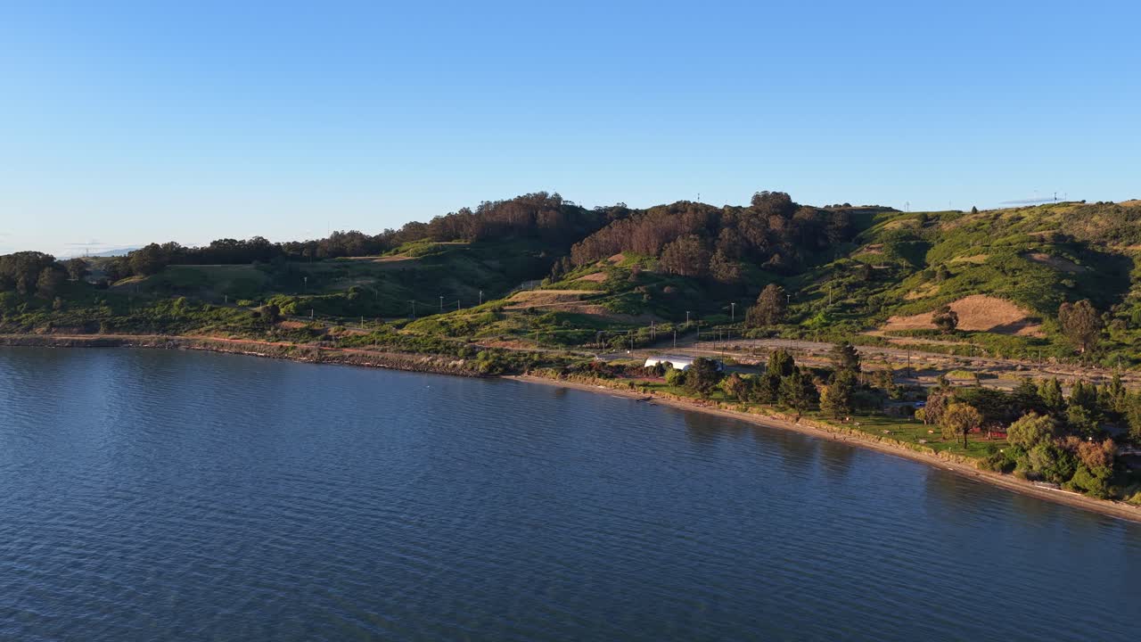 Ripresa aerea con drone che sorvola le calme acque blu della baia di San Francisco, rivelando le dolci colline verdi della costa californiana a Richmond durante un tramonto splendido e limpido