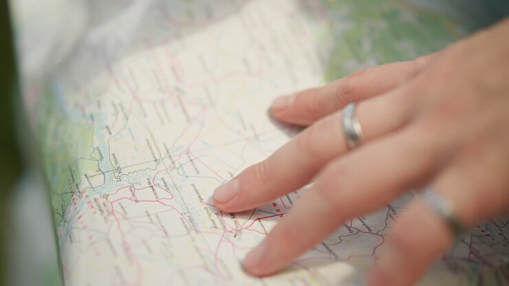 Gros plan sur des mains traçant une carte en papier, lumière extérieure douce, bagues en argent, mouvement de recherche lent, sélection d’itinéraire, planification de randonnée, navigation tactile sur des lignes topographiques, atmosphère de nature sauvage sereine.