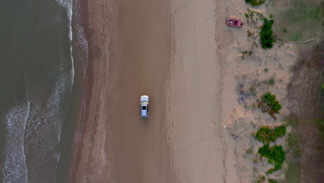 Vista-Aérea-De-Arriba-Hacia-Abajo-Siga-La-Toma-De-Seguimiento-De-Un-Automóvil-Que-Se-Mueve-En-Una-Playa-De-Arena-Cerca-Del-Agua-Durante-El-Anochecer