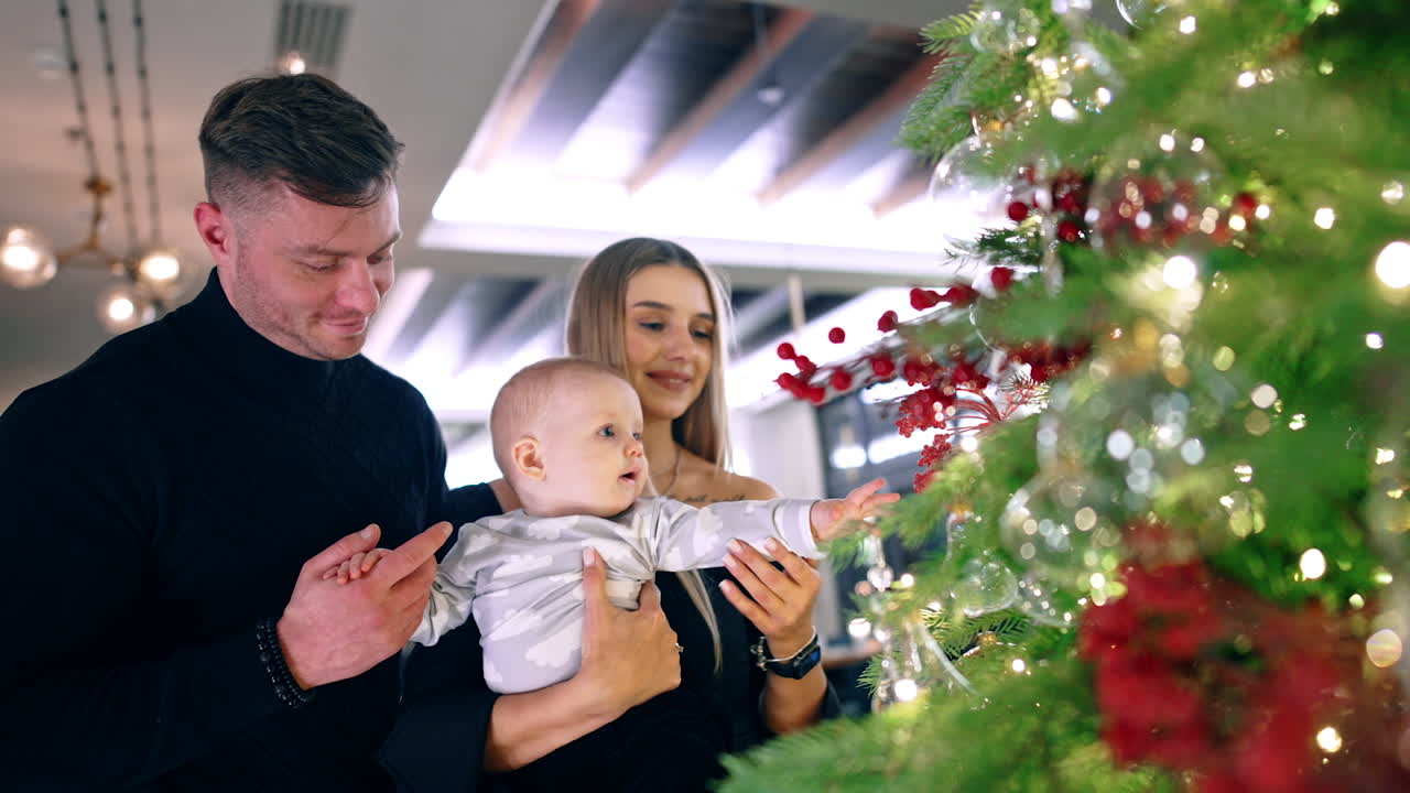 Caucasian family of three standing near the big Christmas tree. Lovely infant boy wants to get the toys.