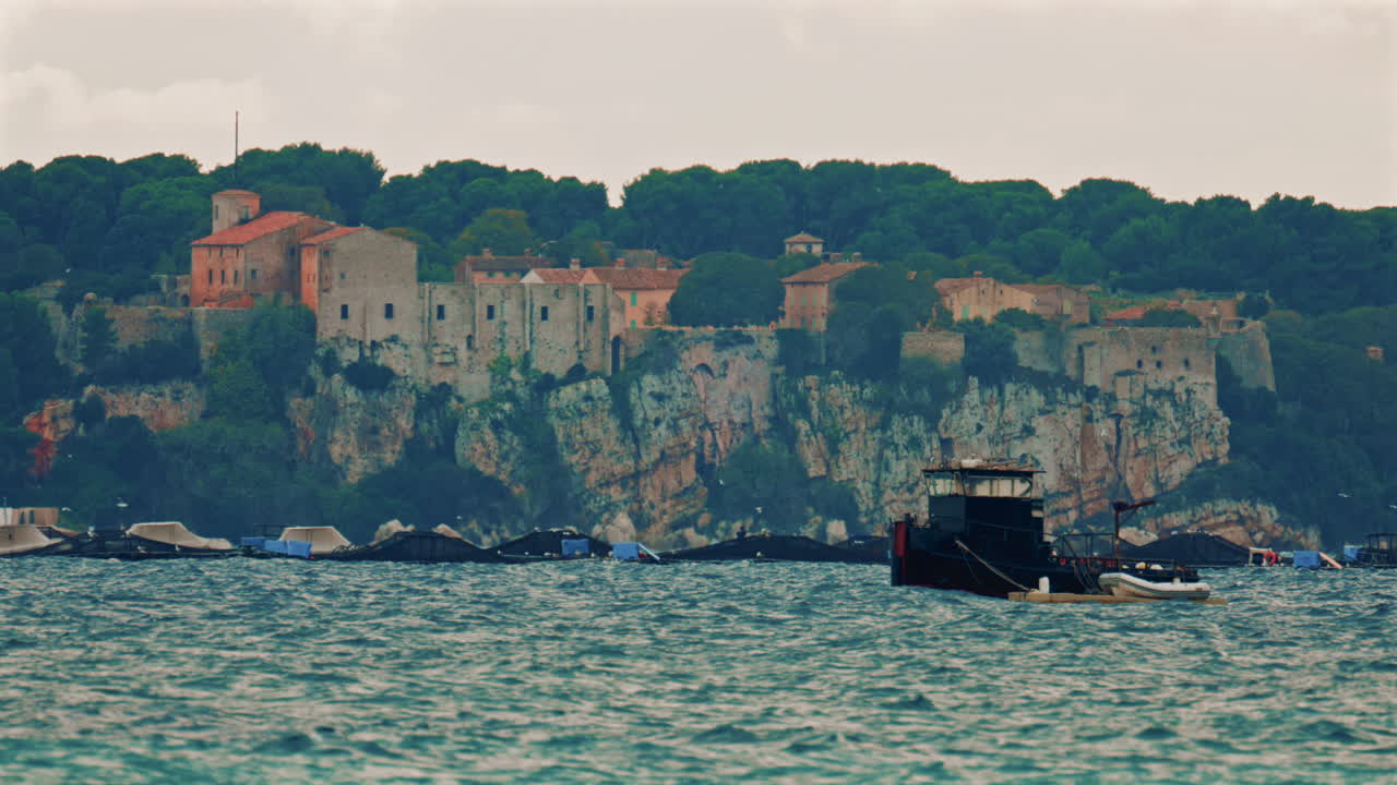 A medieval stone fortress and village perched on a high cliff above the Mediterranean Sea, with a fish farm and moored platforms spread across the water below