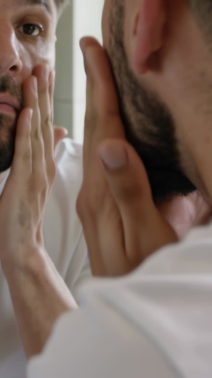 Vertical video: Pushing black pump man in white T rubbing skincare into face, beard at home mirror