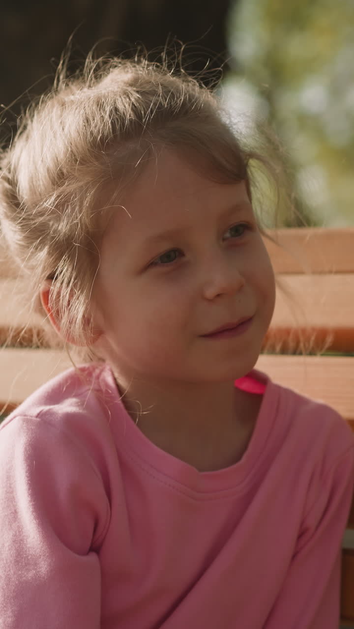 Pretty happy girl sits on wooden bench in sunny park closeup. Cute blonde child spends time in city garden. Positive and healthy kid on spring day