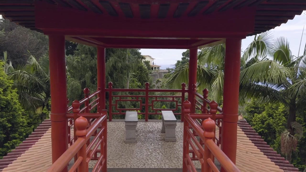 Elegant rød bro i en japansk-inspireret have på Madeira, fra en gående persons synsvinkel mod udsigtsbænke.