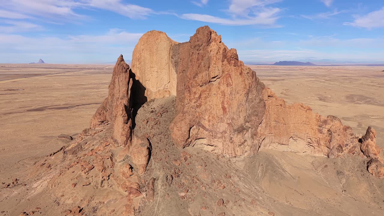 En dramatisk droneflyvning akselererer mot en isolert sandsteinsbutte-monolitt, som viser de robuste geologiske teksturene og den enorme skalaen av en avsidesliggende tørr ørkenvillmark under en blå himmel