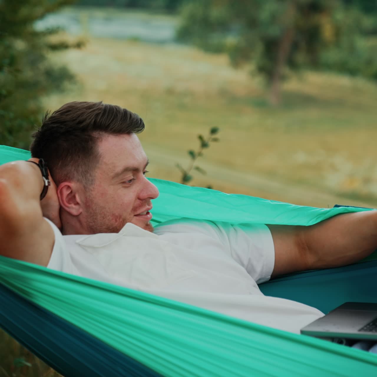 Combining work and pleasure. Caucasian man lies in a hammock and works on laptop. Nature at backdrop