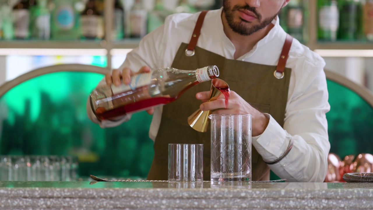 Barman placing mixing glass and cocktail glass. Bartender pouring red bitter and gin with the jigger, working on the counter bar.