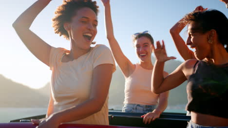 female friends standing up through sun roof of car dancing on road trip