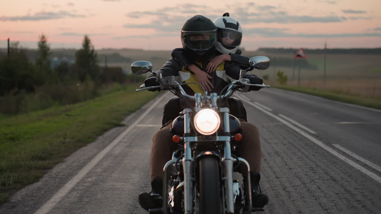 Two friends wearing helmets ride motorcycle on open road at sunset, passenger hugging rider from behind while headlight shines bright, capturing friendship and freedom of countryside travel at dusk