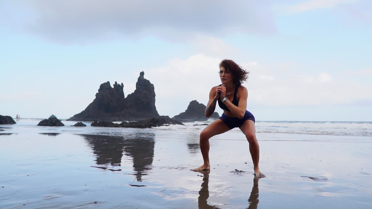 A young, slim woman is seen in slow motion, doing squats and jumps as part of her training by the ocean's shore