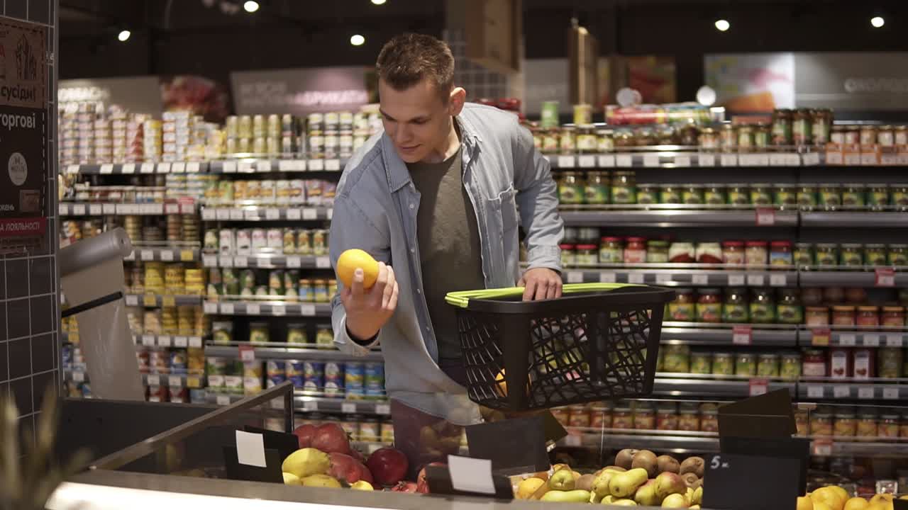 retrato de un hombre joven y positivo comprando frutas y verduras en el departamento de productos de un supermercado de una tienda de comestibles. tomando naranjas y poniéndolas en una canasta negra de transporte manual