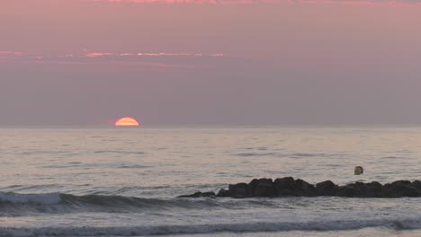 sun rising over ocean horizon into pale dawn skies, waves breaking on rocks