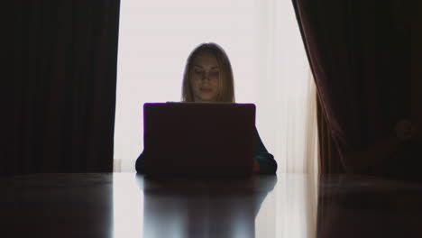 mujer joven abriendo una computadora de cuaderno sentada en la mesa de vuelta a la ventana