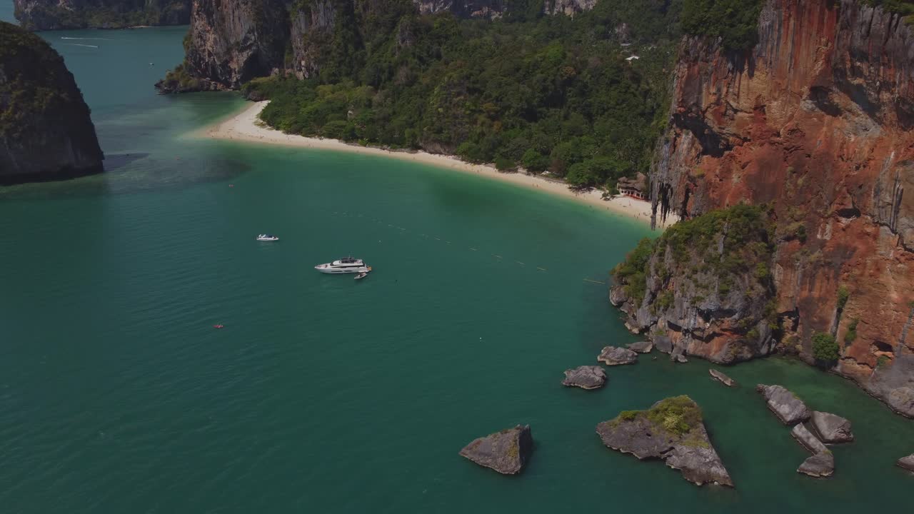 Ripresa aerea della spiaggia di Ao Nang e delle sue spettacolari scogliere calcaree rosse che si ergono sopra il mare turchese, con barche e uno yacht ancorati vicino alla costa tropicale a Krabi, Thailandia