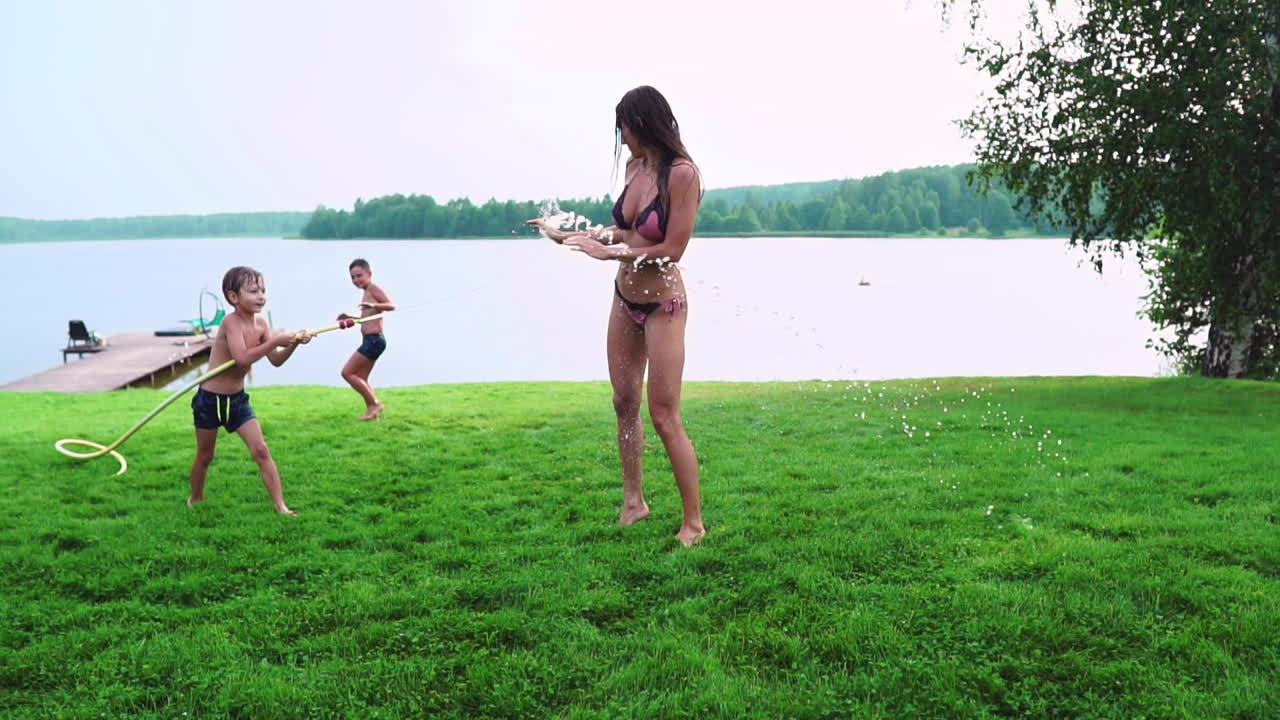 엄마와 아들이 잔디 ⁇ 에서 놀고 물을  ⁇ 고 웃고 놀이터에서 재미를 느끼고 호수 근처의 집의 배경에 잔디 ⁇ 을 가지고 있습니다.