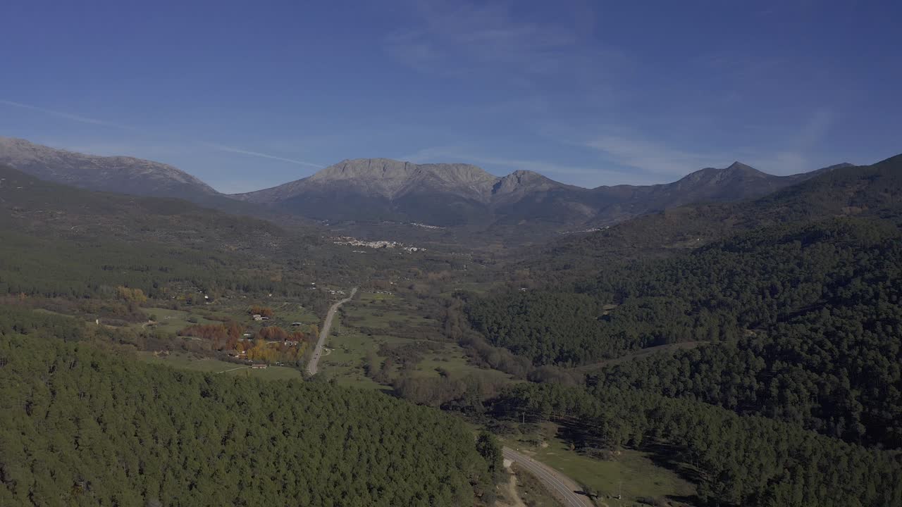 vuelo inverso en un valle con exuberantes bosques de pinos y en el fondo grandes montañas con una carretera con coches en circulación que conduce a un paso de montaña en un día de otoño con en ávila, españa