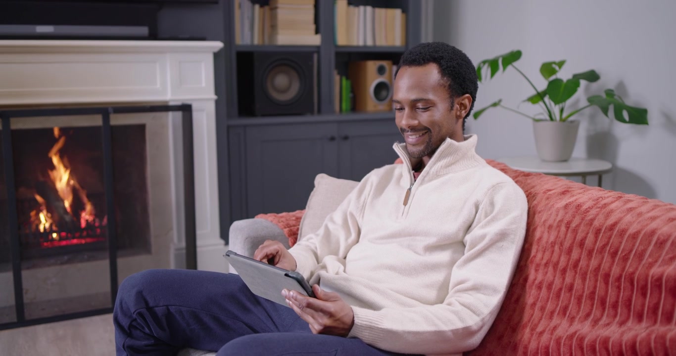 African American man sitting on sofa seeing tablet, tapping, pointing at fire, relaxing in pullover
